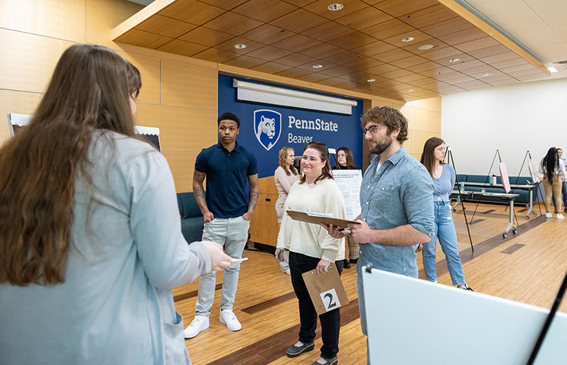 Students and library staff members discuss a student research project. The back wall is blue and displays the Penn State Beaver logo in white.
