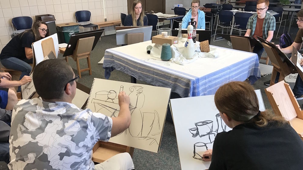Students sit on drawing horses and sketch a still life with charcoal.