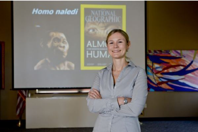 Dr. Heather Garvin poses in front of a screen, projected with imagine of Home naledi.