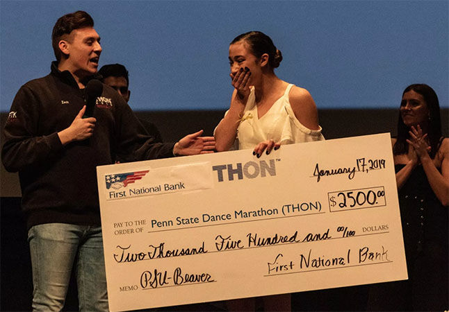 Dancer Hailey Burbage stands on stage, covers her mouth with her hand and reacts with emotion as she is presented a large check for winning the THON Showcase.