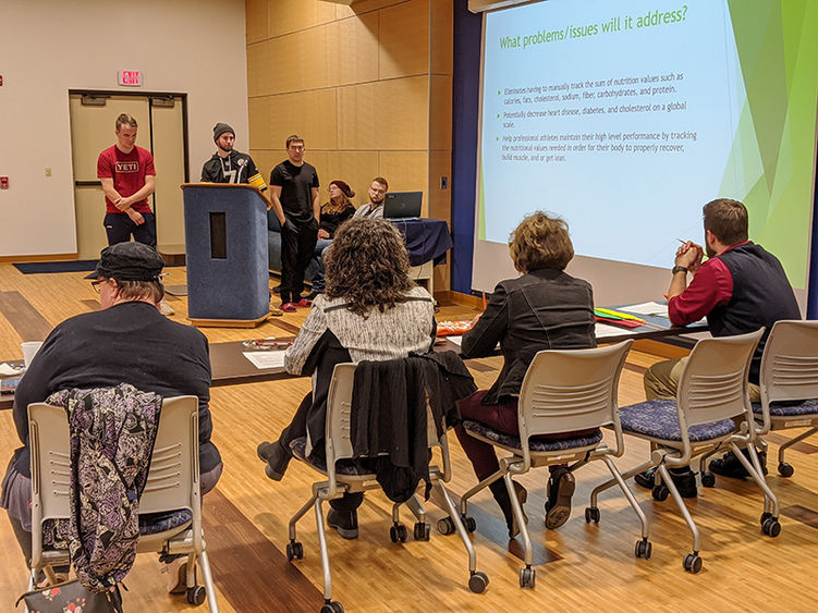 Three male students stand around a podium with a screen projection to the right. A panel of judges is in the foreground of the photo.