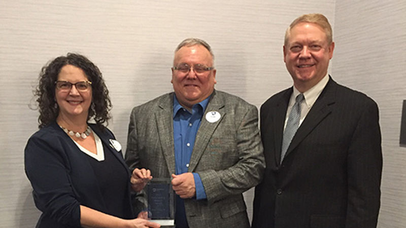 Noxon stands in the center of the photo wearing a blue shirt, brown jacket and holding a glass award. To the left of the photo is Jenifer Cushman wearing a black dress and to the right is Scott Cunningham wearing a dark suit.