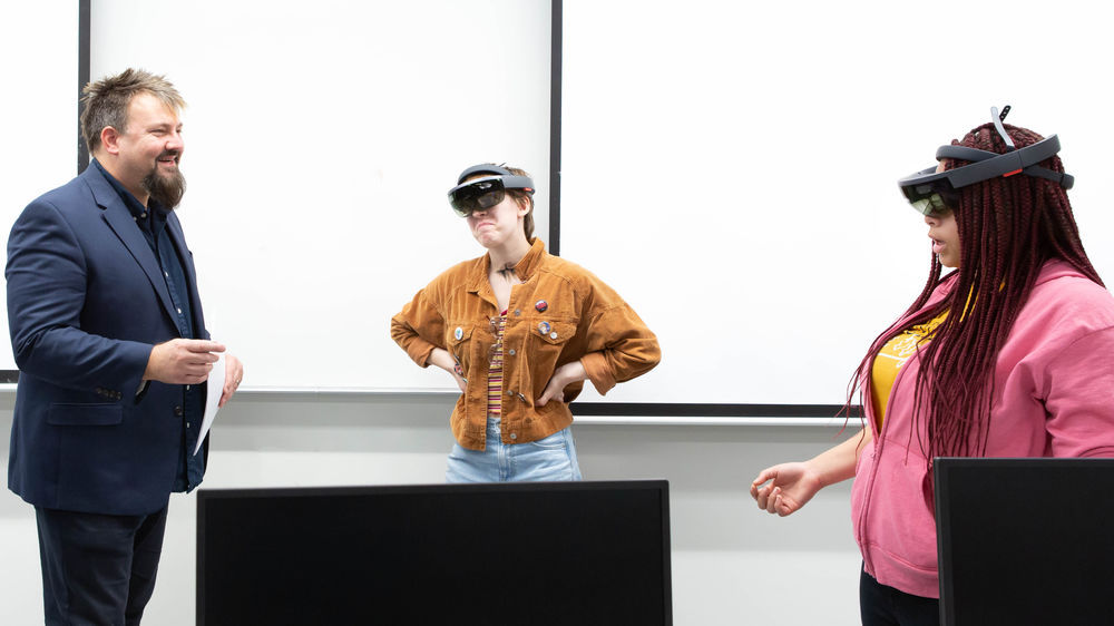 The professor stands to the left as two female students wear goggles as part of a classroom experiment.