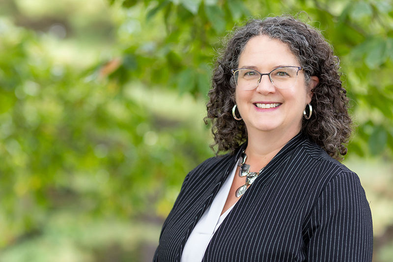 A headshot of Jenifer Cushman standing outdoor in front of a wooded area. She is wearing a dark blazer and white shirt. 