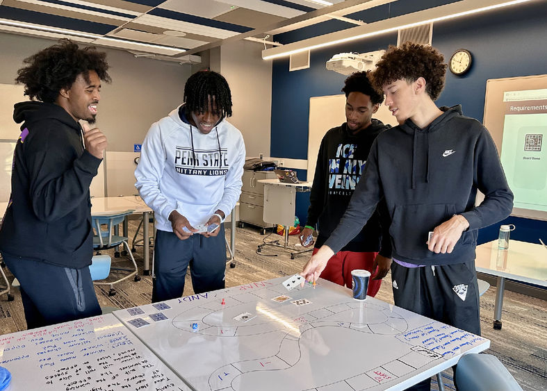 A group of four students stand around a whiteboard table with a custom-made board game on it