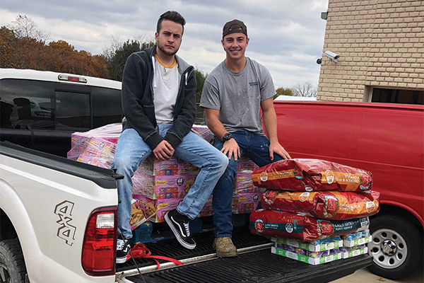 Two students sit on cases of pet food in a pickup truck.