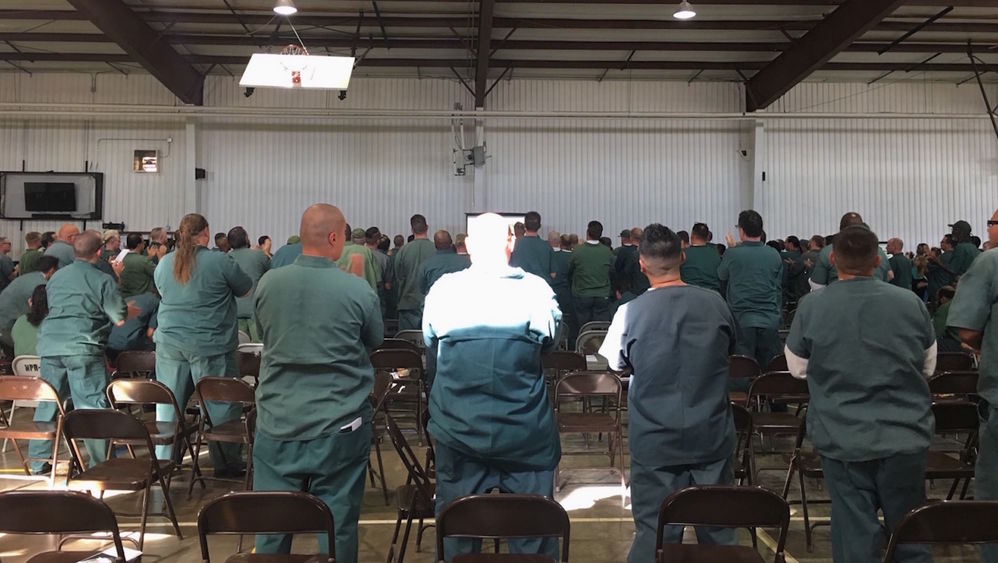 Inmates in Indiana stand in a common room to listen to a speaker.