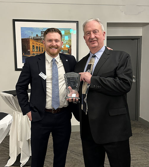 Hertnecky stands at the right of the photo holding a glass award handed to him by Andrew DiPietrantonio who stands at left