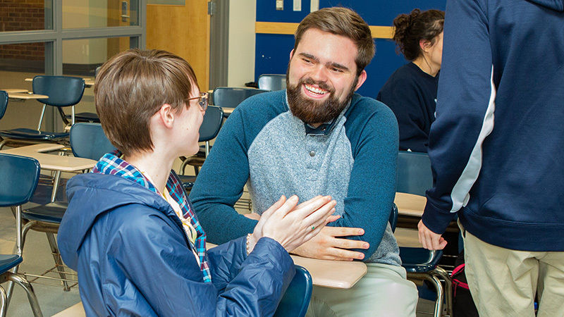 Joshua Hetzer, who has brown hair and a beard and is wearing a blue shirt, is pictured talking to a student during a campus activity.