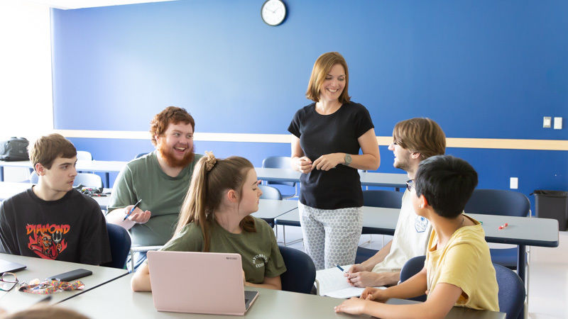 A female professor in a black t-shirt stands and interacts with a group of seated students