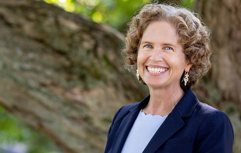 McDougall is pictured outdoors with trees in the background. She is smiling and wearing a light blue shirt and navy blue blazer.