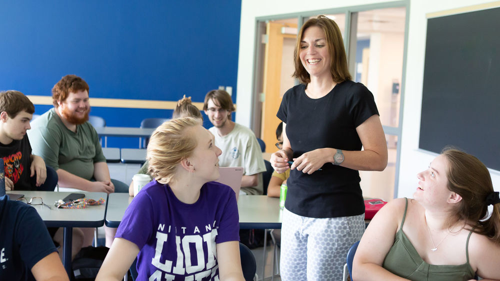 A woman in a black shirt stands smiling as she teaches a group of students who are seated around her.