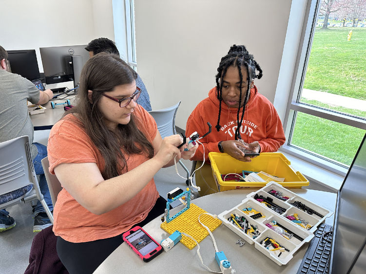 Stephanie wearing a salmon colored shirt and Tyera wearing an orange shirt work in a classroom on their builds