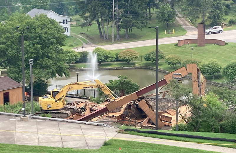 A piece of construction equipment sits in front of the remnants of a building. There is a pond with a fountain in the background.